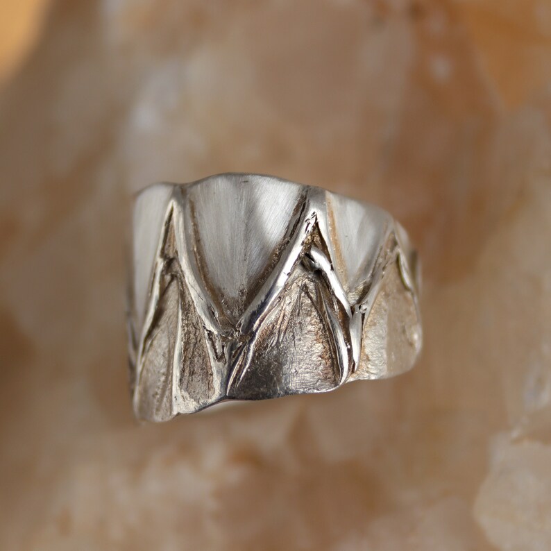 May include: A silver ring with a textured design of overlapping triangles. The ring is polished and has a shiny finish.