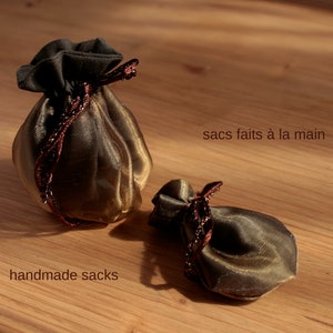 May include: Two small, handmade, gold-colored sacks with brown ribbon ties. The sacks are made of a sheer fabric and are sitting on a wooden surface. The text "handmade sacks" is written below the sacks, and the text "sacs faits &agrave; la main" is written above the sacks.