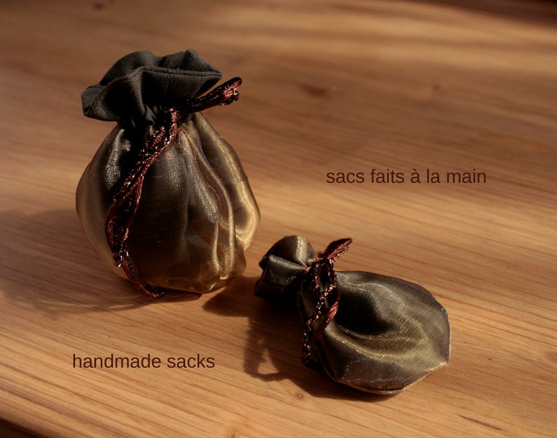 May include: Two small, handmade, gold-colored sacks with brown ribbon ties. The sacks are made of a sheer fabric and are sitting on a wooden surface. The text "handmade sacks" is written below the sacks, and the text "sacs faits &agrave; la main" is written above the sacks.