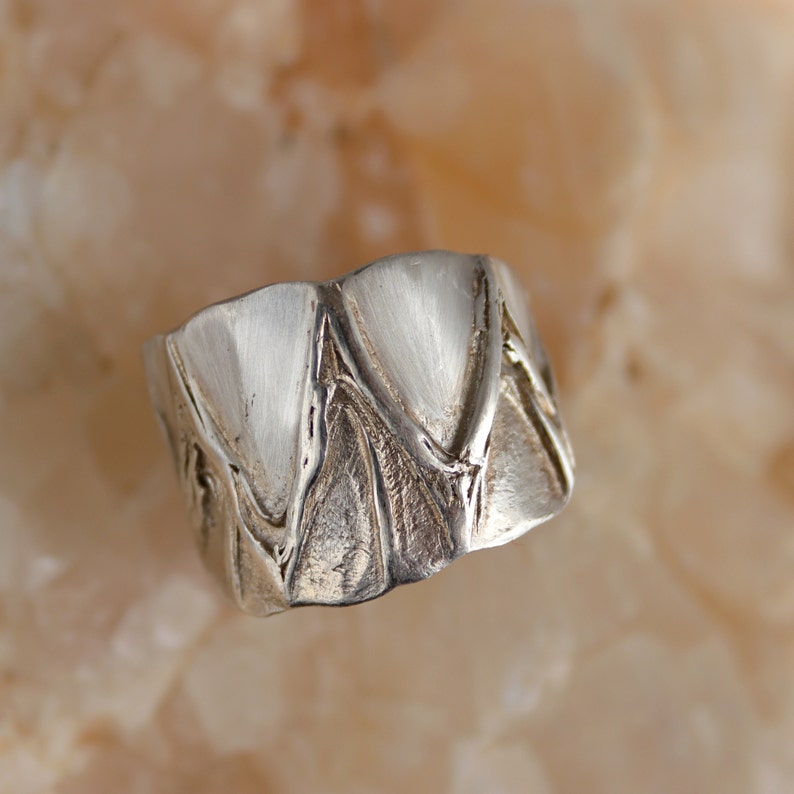 May include: A silver ring with a textured, geometric design. The ring is made of metal and has a shiny finish.