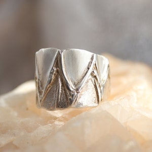 May include: A silver ring with a textured, geometric design. The ring is made of a smooth, polished metal and has a unique, modern look.