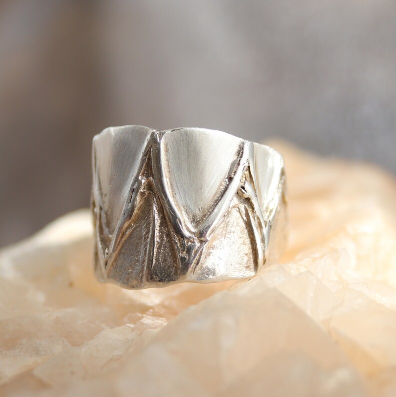 May include: A silver ring with a textured, geometric design. The ring is made of a smooth, polished metal and has a unique, modern look.
