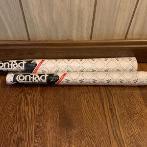 May include: Two rolls of self-adhesive contact paper. The rolls are white with a repeating pattern of red and green floral designs. The brand name "On-Tact" is printed on the end of each roll in black and white.