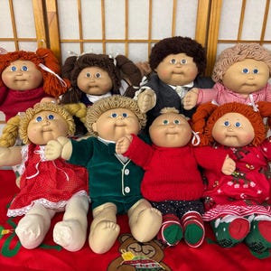 May include: A collection of eight Cabbage Patch Kids dolls with yarn hair, soft bodies, and vinyl faces. The dolls wear various outfits including red dresses, a green velvet suit, a red sweater, and patterned shirts, all featuring blue eyes.