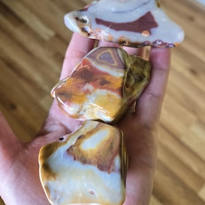 Puede incluir: Tres piedras pulidas con patrones y colores naturales. La piedra superior es blanca con marcas granates y marrones. La piedra del medio es una mezcla de naranja, amarillo y marrón. La piedra inferior es blanca, marrón y amarilla. Estas piedras son ideales para coleccionistas o decoración.