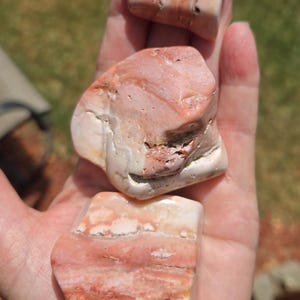 May include: Three polished tumbled stones with a smooth texture, featuring a color palette of soft pink, creamy white, and deep reddish-brown bands. The stones have irregular, rounded shapes and a glossy finish that reflects light across their surfaces.