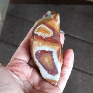 Puede incluir: Una piedra de ágata lisa y pulida, de color marrón, blanco y naranja, con un patrón único en espiral. La piedra tiene forma de corazón y una superficie lisa y pulida.