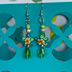 May include: A pair of green and floral dangle earrings with green crystal drops. The earrings are made with silver hooks and feature a green and yellow bead at the top.