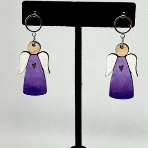 May include: A pair of wooden angel earrings. Each earring features a purple and white angel design with a small heart detail. The earrings hang from silver-colored hoops.