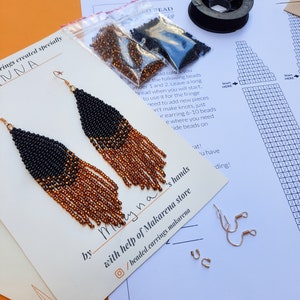 May include: A pair of black and orange beaded fringe earrings with gold ear wires. The earrings are displayed on a white card with the text "Maryna" and "with help of Makarena store" written on it. The card also includes a social media icon and the text "/beaded.earrings.makarena".