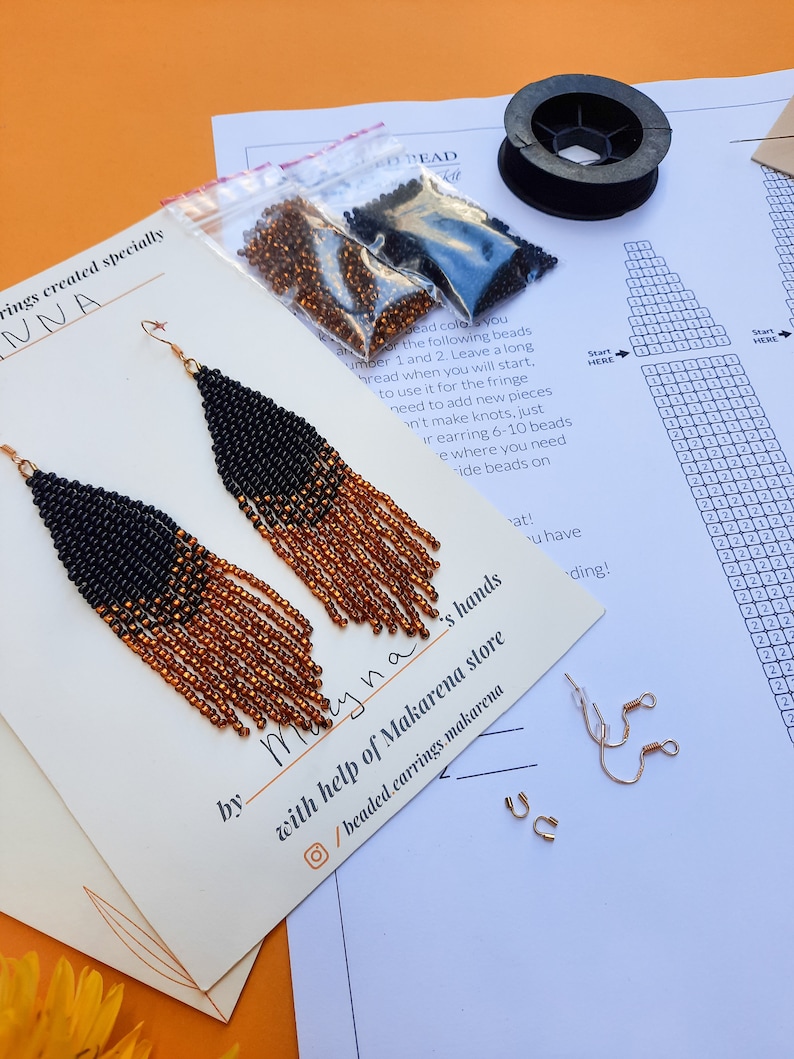 May include: A pair of black and orange beaded fringe earrings with gold ear wires. The earrings are displayed on a white card with the text "Maryna" and "with help of Makarena store" written on it. The card also includes a social media icon and the text "/beaded.earrings.makarena".