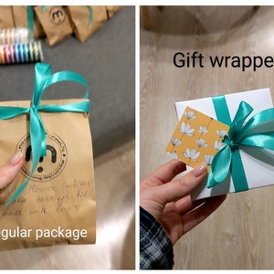 May include: Two images showing packaging options for a set of earrings. The first image shows a brown paper bag with a teal ribbon tied around it. The bag has a handwritten note that says "flower (white) beaded earrings kit made with love". The second image shows a white gift box with a teal ribbon tied around it. The box has a small yellow tag with white flowers on it.