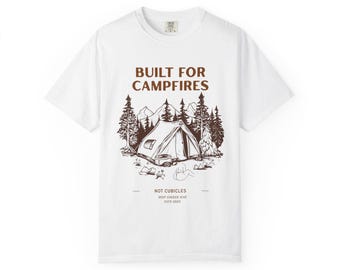 May include: White t-shirt with a brown graphic of a tent, trees, and mountains. The text "BUILT FOR CAMPFIRES" is above the graphic. Below the graphic, the text reads "NOT CUBICLES, DEEP CHECK DIVE, ESTD 2025".