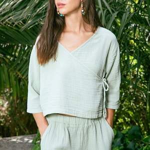 May include: A light green, loose-fitting, wrap-style top with a V-neck and 3/4 sleeves. The top is paired with matching light green trousers. The model is standing in front of a lush green background.