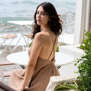 May include: A woman wearing a brown sleeveless dress with a criss-cross back detail sits on a white bench by the ocean.