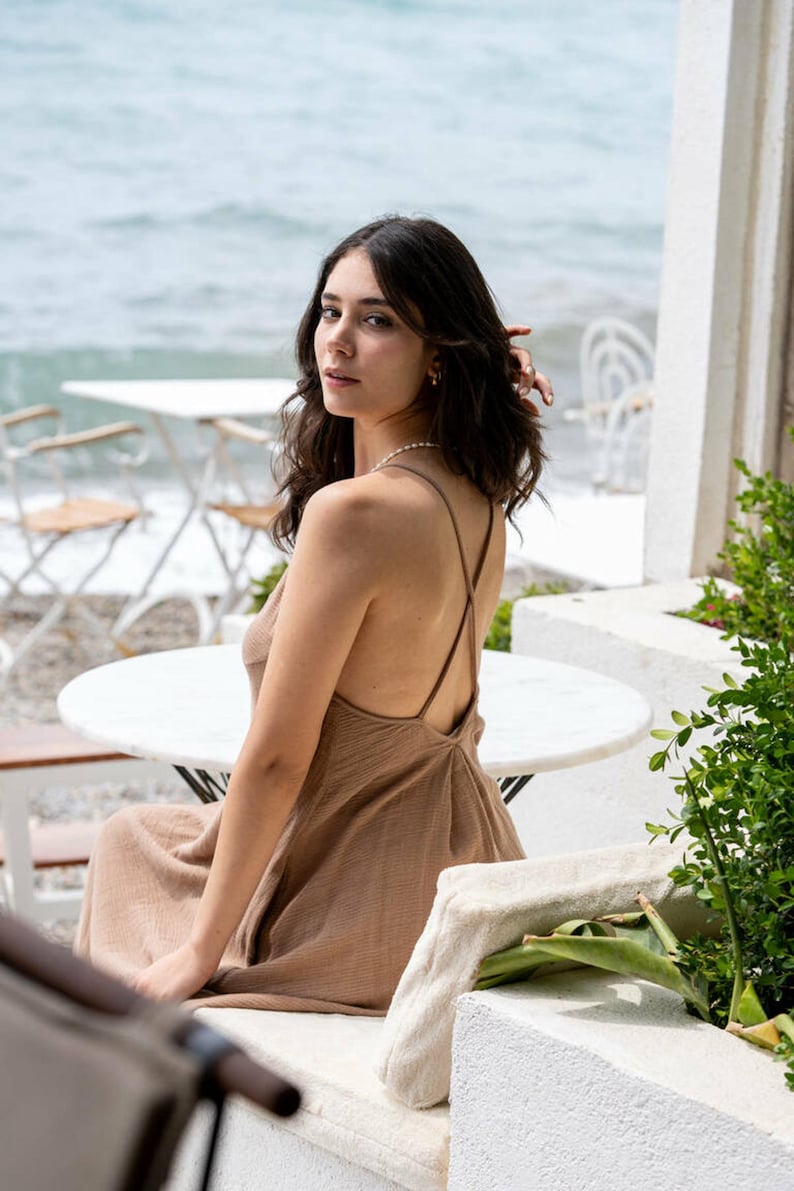 May include: A woman wearing a brown sleeveless dress with a criss-cross back detail sits on a white bench by the ocean.