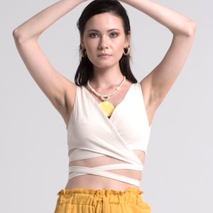 May include: A woman in a cream wrap top and mustard yellow trousers. The top features a crisscross design, and the trousers are high-waisted. She wears gold hoop earrings and a necklace with a yellow pendant. The outfit is set against a white backdrop.