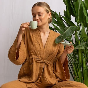 May include: A person wearing a brown, loose-fitting, long-sleeved top with a drawstring waist and matching trousers. The person is sitting on a white surface and holding a light green cup and saucer.