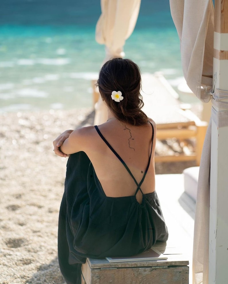 May include: A woman wearing a black backless dress with a white flower in her hair sits on a wooden bench facing the ocean.