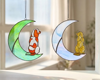 May include: Two stained glass suncatchers hanging by black cords. One features a green crescent moon with a white and orange dog, while the other shows a light blue crescent moon with a golden-yellow dog sitting on the curve.