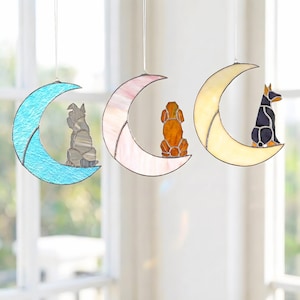 May include: Three stained glass dog suncatchers. Each dog is perched on a crescent moon. The moons are blue, pink, and yellow. The dogs are gray, orange, and black. The suncatchers hang from clear strings.