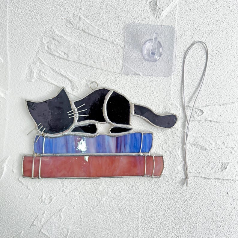 May include: A stained glass cat figurine, black with white whiskers, is lying on top of a stack of three books. The books are blue, purple, and red. The cat is attached to a silver string.