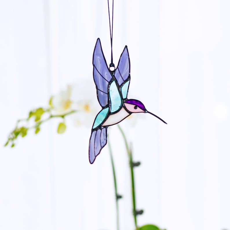 May include: A stained glass hummingbird suncatcher with purple, blue, and pink accents. The hummingbird is in flight with its wings spread out. The suncatcher is hanging from a black string.