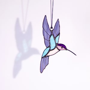 May include: A stained glass hummingbird ornament with purple, blue, and pink accents. The hummingbird is in flight with its wings spread out.