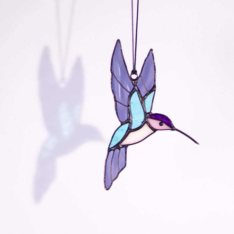 May include: A stained glass hummingbird ornament with purple, blue, and pink accents. The hummingbird is in flight with its wings spread out.