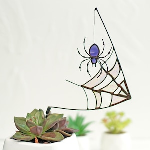 May include: A decorative Halloween spider and web hanging ornament. The spider has a purple body and black legs, suspended from a black wire. The web is a translucent, iridescent material. The ornament is set against a white background.