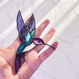 May include: A stained glass hummingbird with purple, blue, and white wings. The hummingbird is in flight with its beak extended.