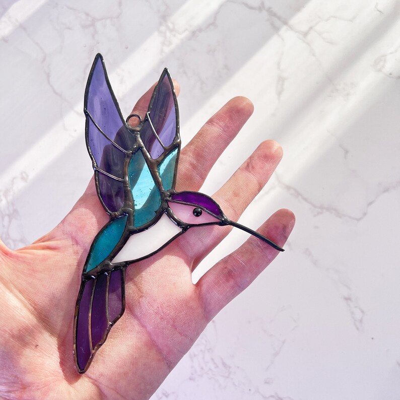 May include: A stained glass hummingbird with purple, blue, and white wings. The hummingbird is in flight with its beak extended.