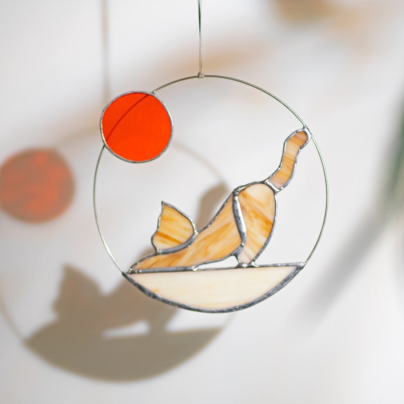 May include: A stained glass suncatcher featuring a cat in a yoga pose with a red sun in the background.