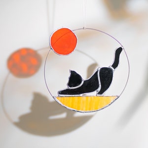 May include: A black cat stained glass suncatcher hanging from a wire with a yellow and orange background. The cat is in a yoga pose with its back arched and its head down. The suncatcher is surrounded by a clear circle.