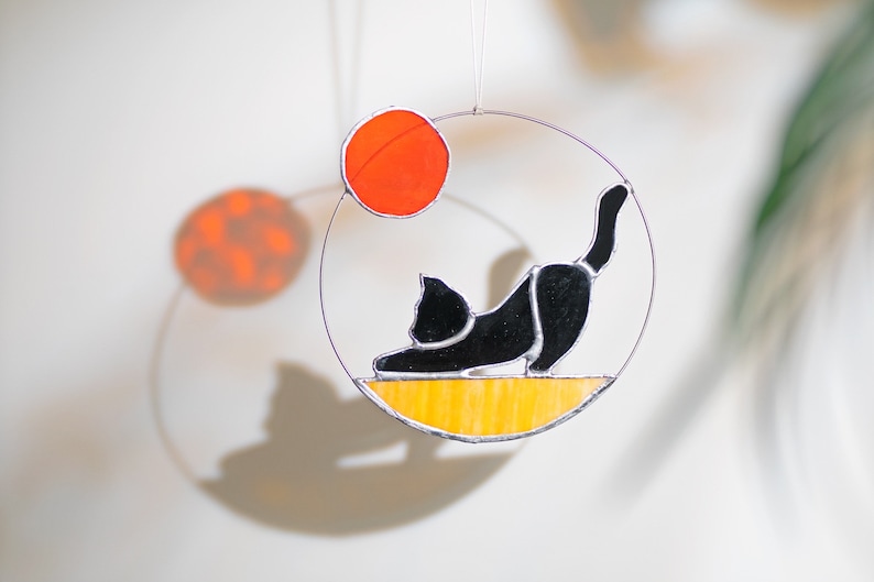 May include: A black cat stained glass suncatcher hanging from a wire with a yellow and orange background. The cat is in a yoga pose with its back arched and its head down. The suncatcher is surrounded by a clear circle.