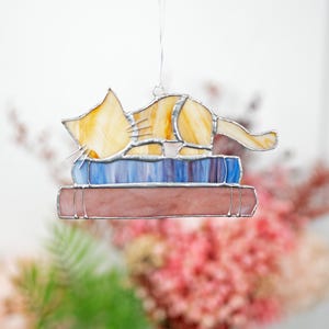 May include: A stained glass cat figurine sleeping on a stack of books. The cat is yellow and orange with a blue and purple book underneath a pink book.