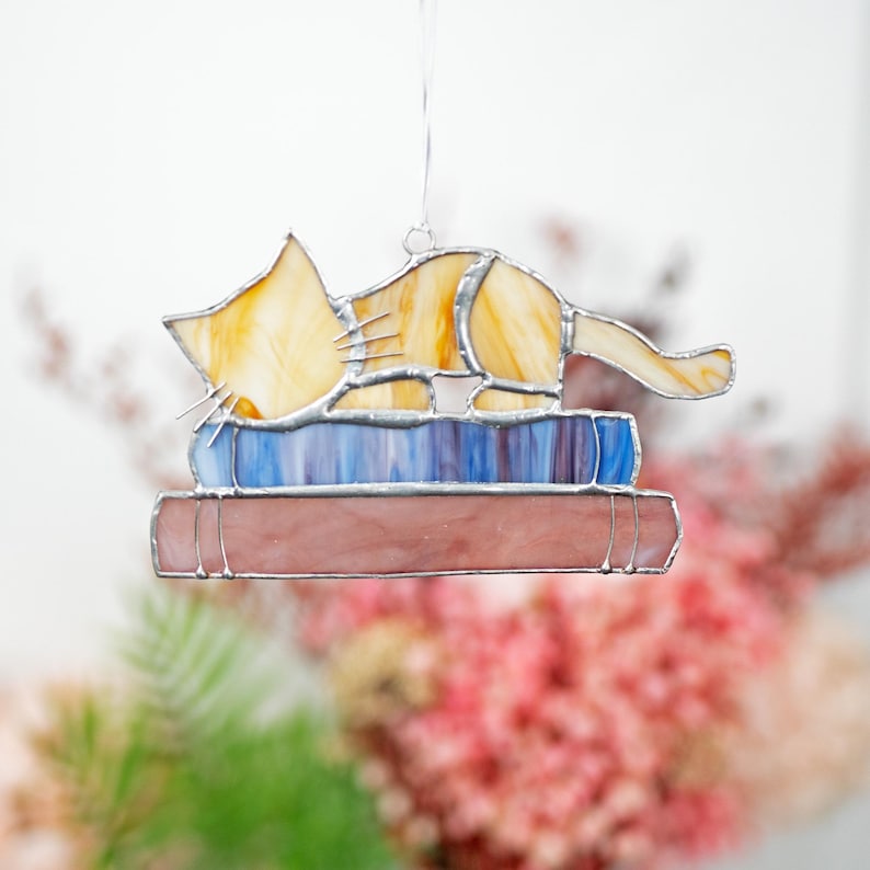May include: A stained glass cat figurine sleeping on a stack of books. The cat is yellow and orange with a blue and purple book underneath a pink book.