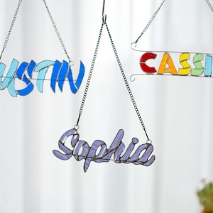 May include: Three stained glass name signs hanging from chains. The names are Justin, Cassie, and Sophia. The Justin sign is blue, the Cassie sign is rainbow, and the Sophia sign is purple.
