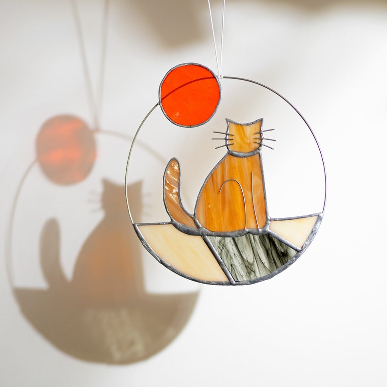 May include: A stained glass suncatcher featuring a cat sitting on a rock with a red sun in the background.