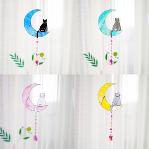 May include: Four stained glass moon and cat suncatchers with crystal beads hanging from the bottom. The moons are blue, pink, yellow, and green. The cats are black, white, gray, and gray. The cats are sitting on the crescent moons. The suncatchers are hanging from a white background.