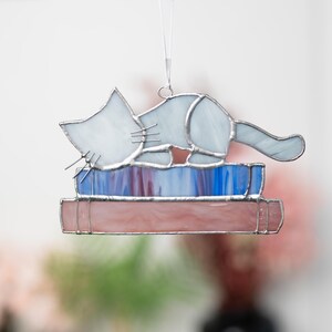 May include: A white stained glass cat figurine laying on top of two stacked stained glass books. The books are pink, blue, and purple.