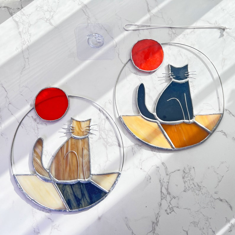 May include: Two stained glass cat suncatchers, one with a black cat and the other with a brown cat. Both cats are sitting and facing the right. The suncatchers are round and have a red circle in the top half. The background is a white marble surface.