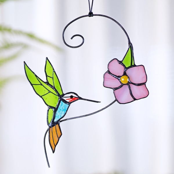 May include: A stained glass hummingbird with green, blue, and orange wings, perched on a black metal branch with a pink flower. The hummingbird is facing right and has a red beak. The flower has a yellow center.