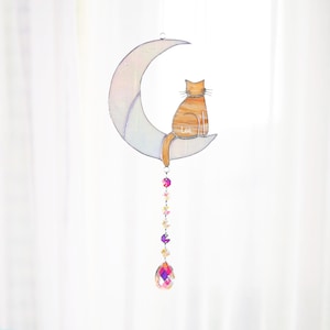 May include: A stained glass moon with a cat sitting on it. The cat is orange and white and has the name "Lee" written on it. The moon is iridescent and has a crystal hanging from it.