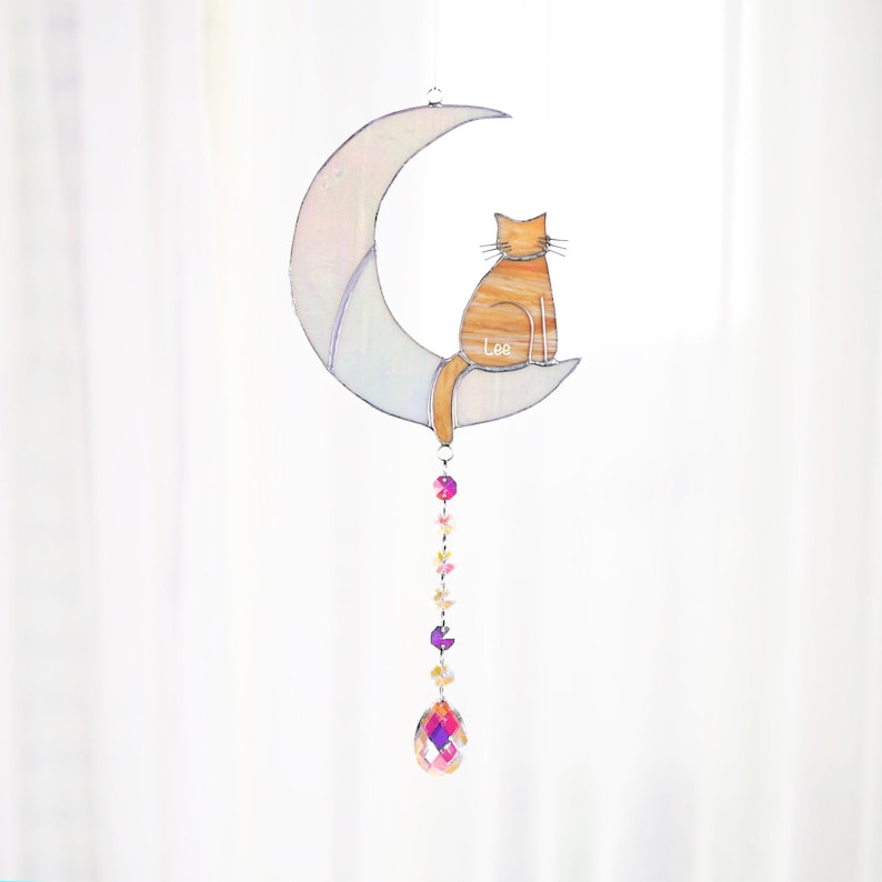 May include: A stained glass moon with a cat sitting on it. The cat is orange and white and has the name "Lee" written on it. The moon is iridescent and has a crystal hanging from it.