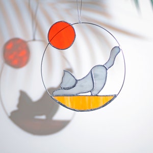 May include: A stained glass suncatcher featuring a gray cat in a yoga pose on a yellow and orange background. The cat is facing left and has a white circle around it. The suncatcher is hanging from a silver wire.