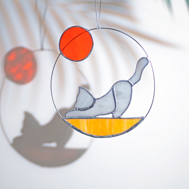 May include: A stained glass suncatcher featuring a gray cat in a yoga pose on a yellow and orange background. The cat is facing left and has a white circle around it. The suncatcher is hanging from a silver wire.