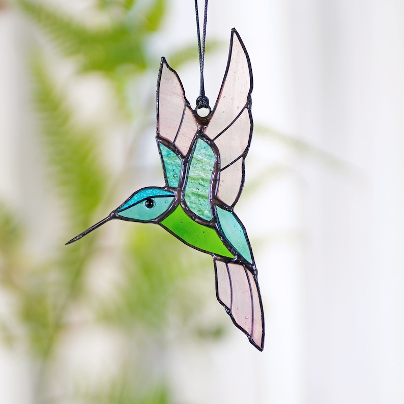 May include: A stained glass hummingbird ornament with pink, green, and blue accents. The hummingbird is in flight with its wings spread out.