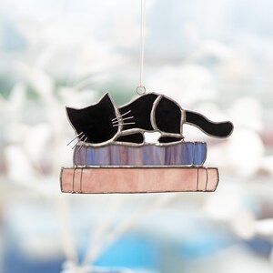 May include: A black cat figurine sleeping on a stack of colorful books. The cat is made of stained glass and has a detailed design. The books are also made of stained glass and have a variety of colors, including pink, blue, and purple.