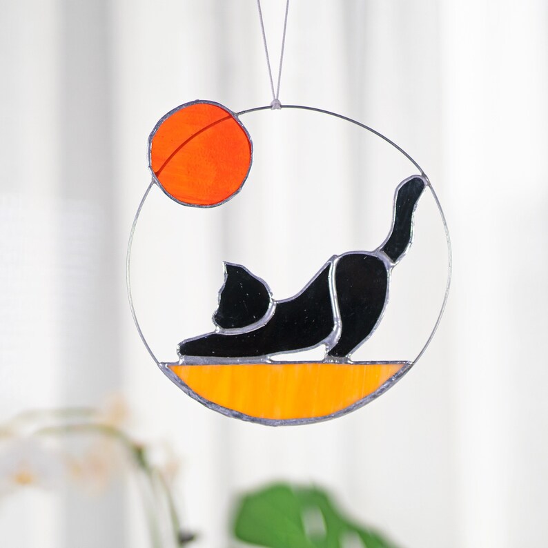 May include: A stained glass suncatcher featuring a black cat stretching with an orange circle above it.